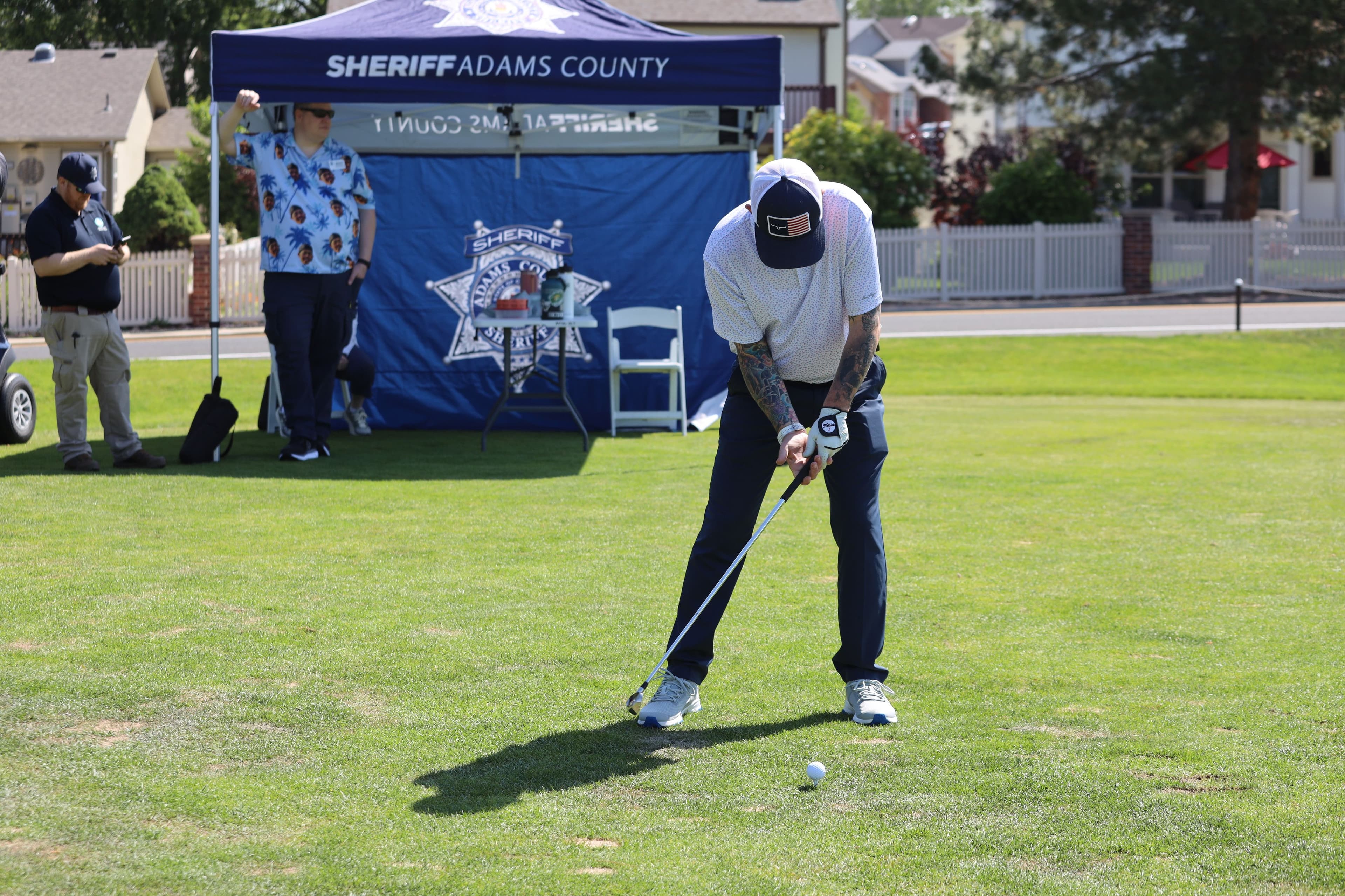 A golfer tees off during the Sheriff's Office Employees' Orphans Fund Golf Tournament, with an Adams County Sheriff's Office event tent in the background.