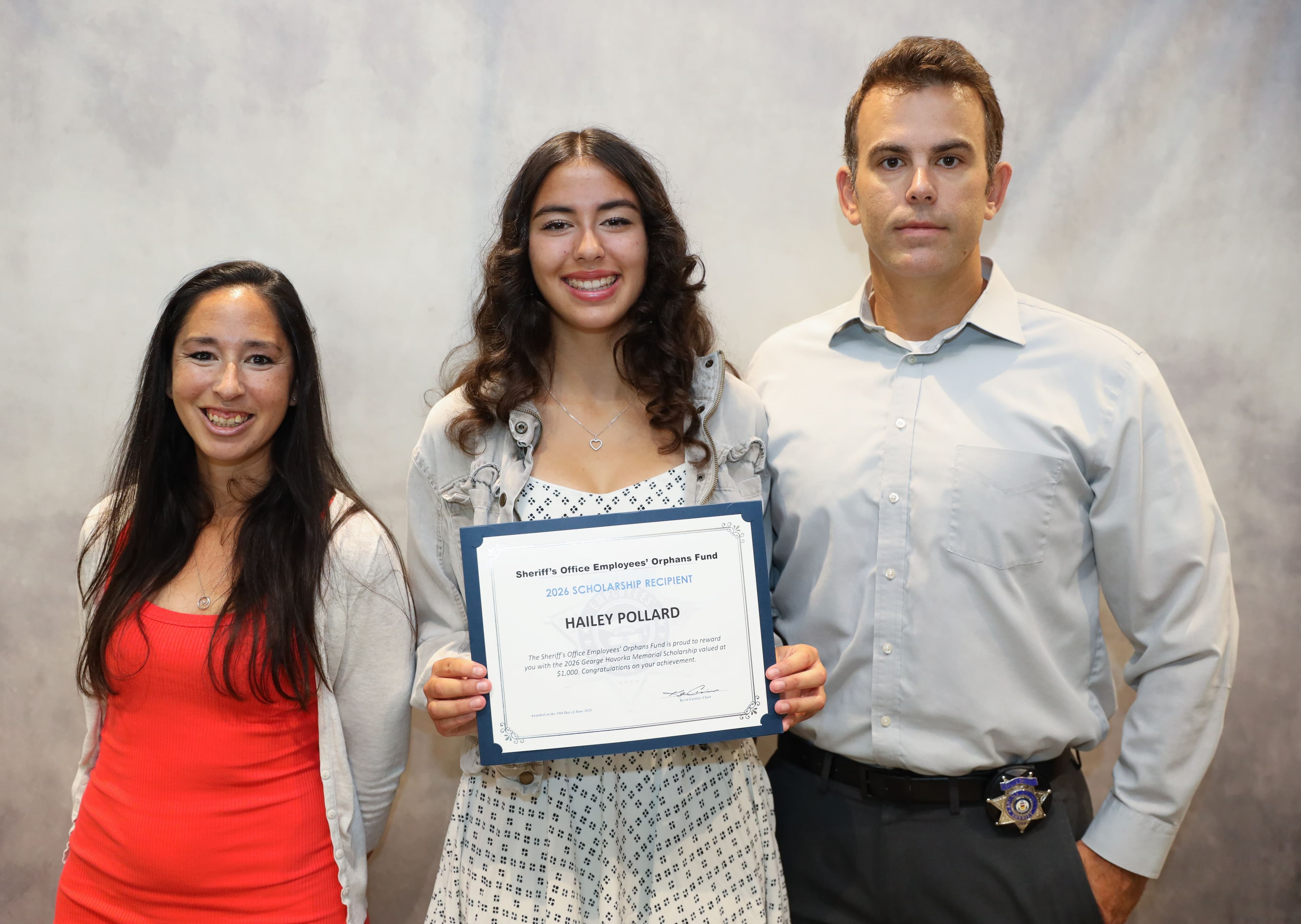 Hailey Pollard smiles while holding her 2026 Scholarship Recipient certificate, standing between her parents against a grey background.