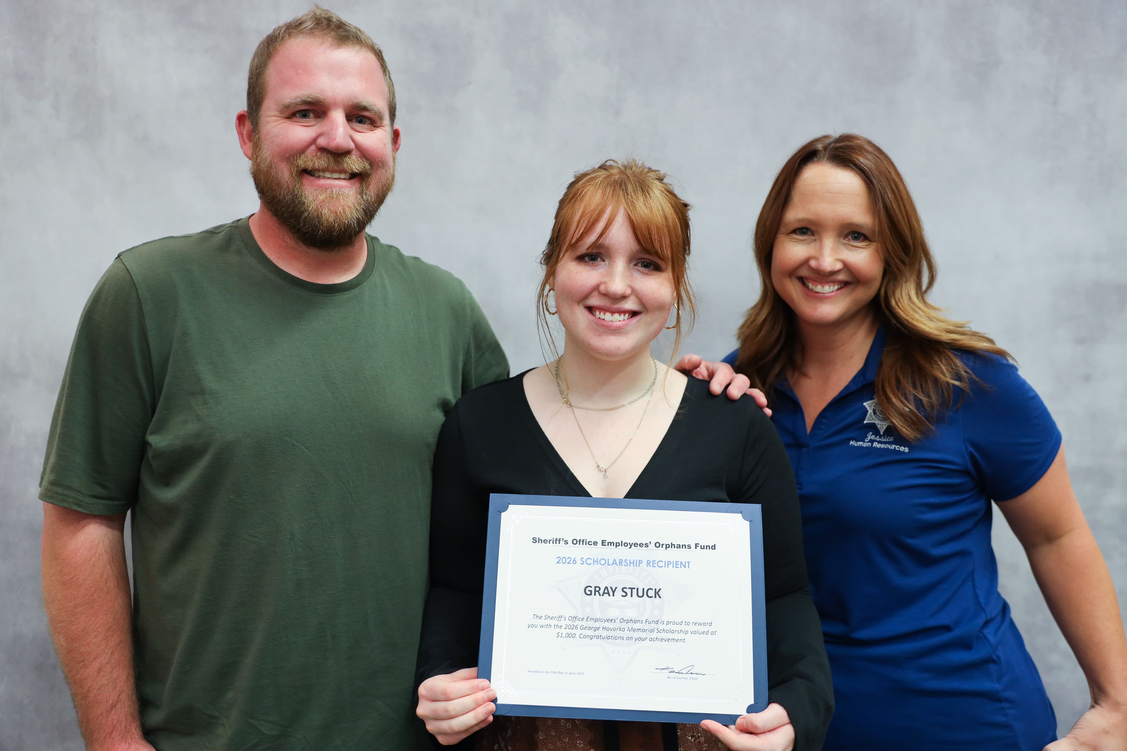 Gray Stuck smiles while holding her 2026 Scholarship Recipient certificate, standing between her parents against a grey background.
