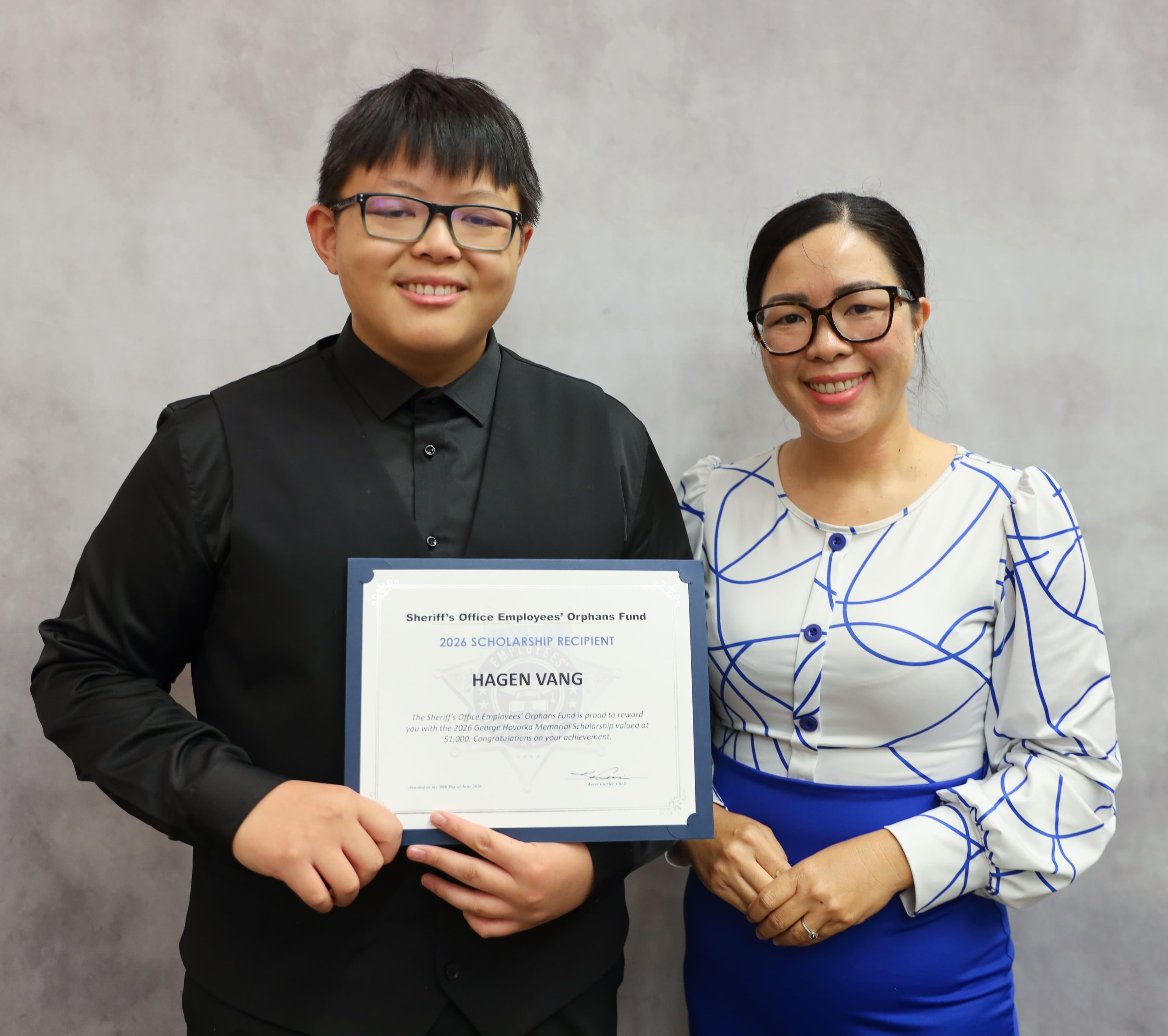 Hagen Vang smiles while holding a 2026 Scholarship Recipient certificate next to his mother against a grey background.